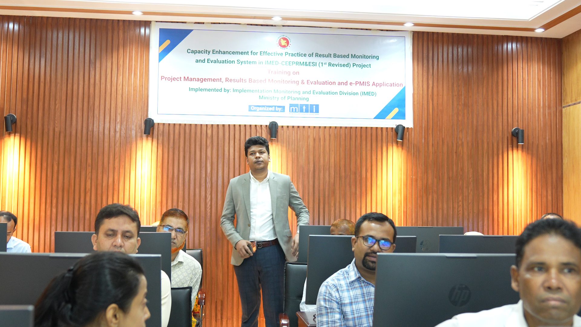 Raihan Khan addresses seated participants in the computer lab, standing beneath the project's training banner.