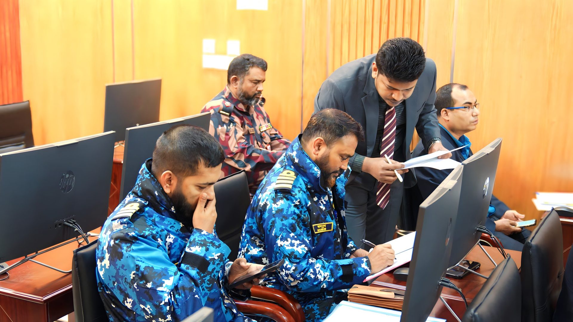 Raihan Khan in a grey suit leans in to assist two Bangladesh Navy officers at their computer terminals during a training session.