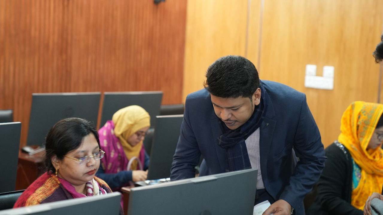 Raihan Khan in a navy blazer helps a seated woman participant at her computer as colleagues in colorful hijabs work at nearby desks.