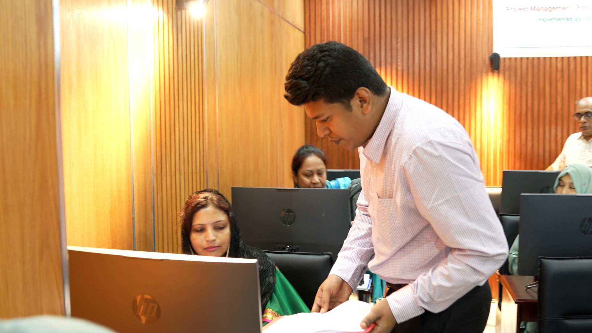 A facilitator in a light pink shirt leans over to guide a seated woman participant at her computer during a training batch session.