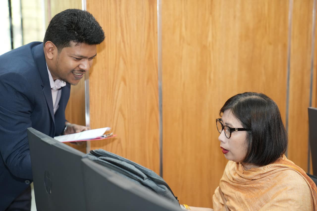 Raihan Khan smiles while briefing a woman participant in an orange shawl at her computer workstation.