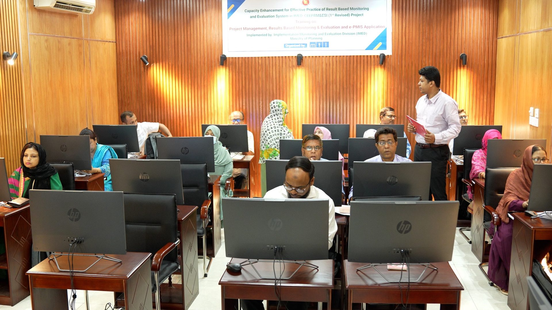 A full classroom of officials attends the training session as a facilitator walks the rows holding a folder.