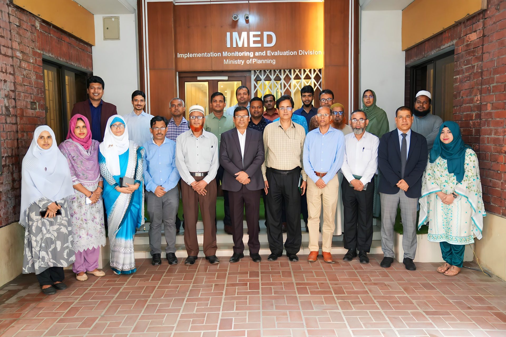 A large group of training participants and IMED officials, including a Bangladesh Navy officer and a police officer, pose together in formal business attire beneath the IMED signage.