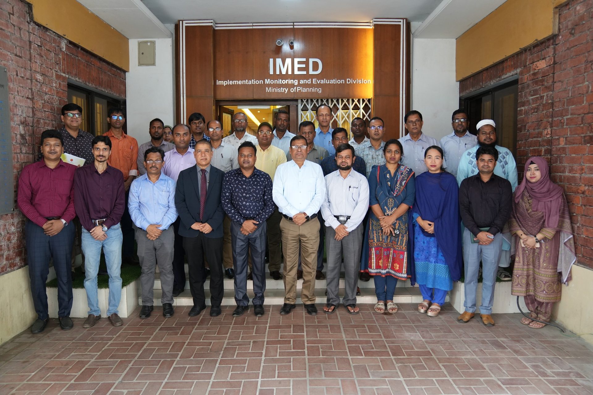 Group photo of training participants beneath the IMED signage, with a woman in a blue and gold traditional dress standing among colleagues in business attire.