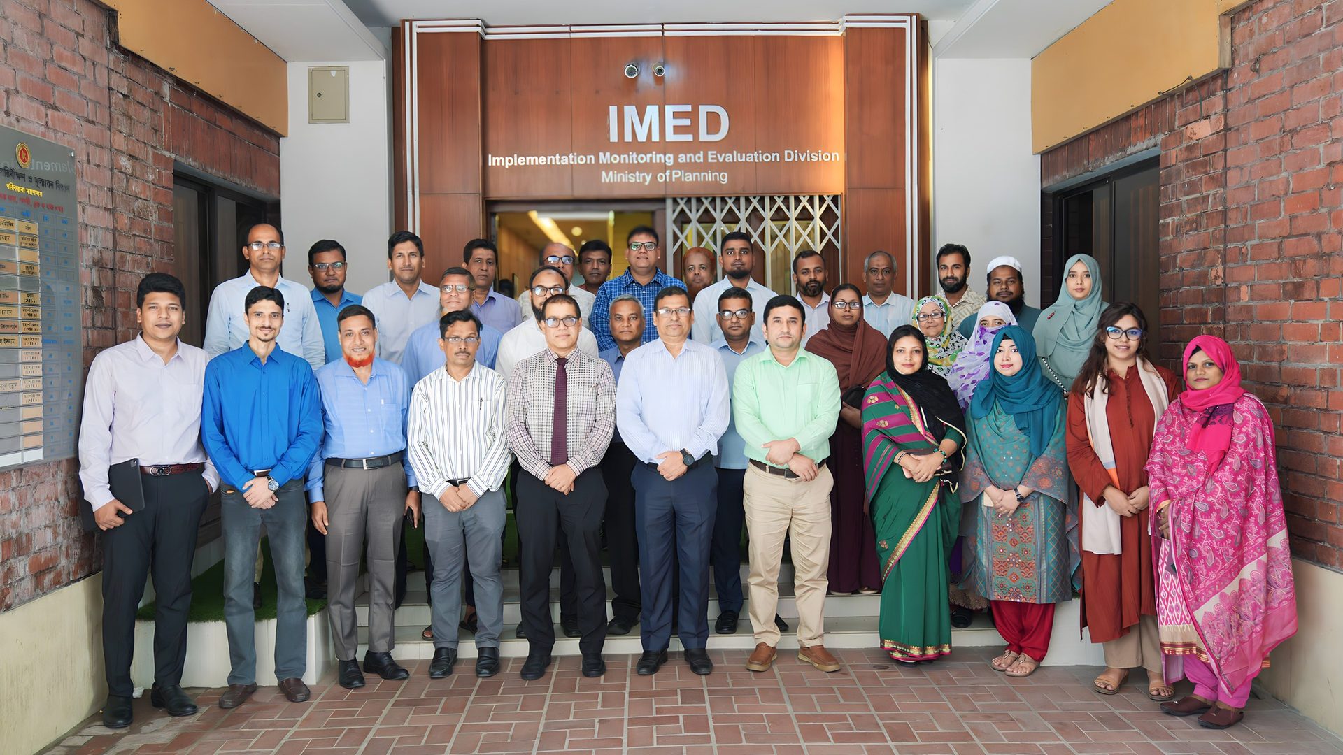 Group photo of training participants under the IMED signage, featuring a woman in a golden saree in the front row.