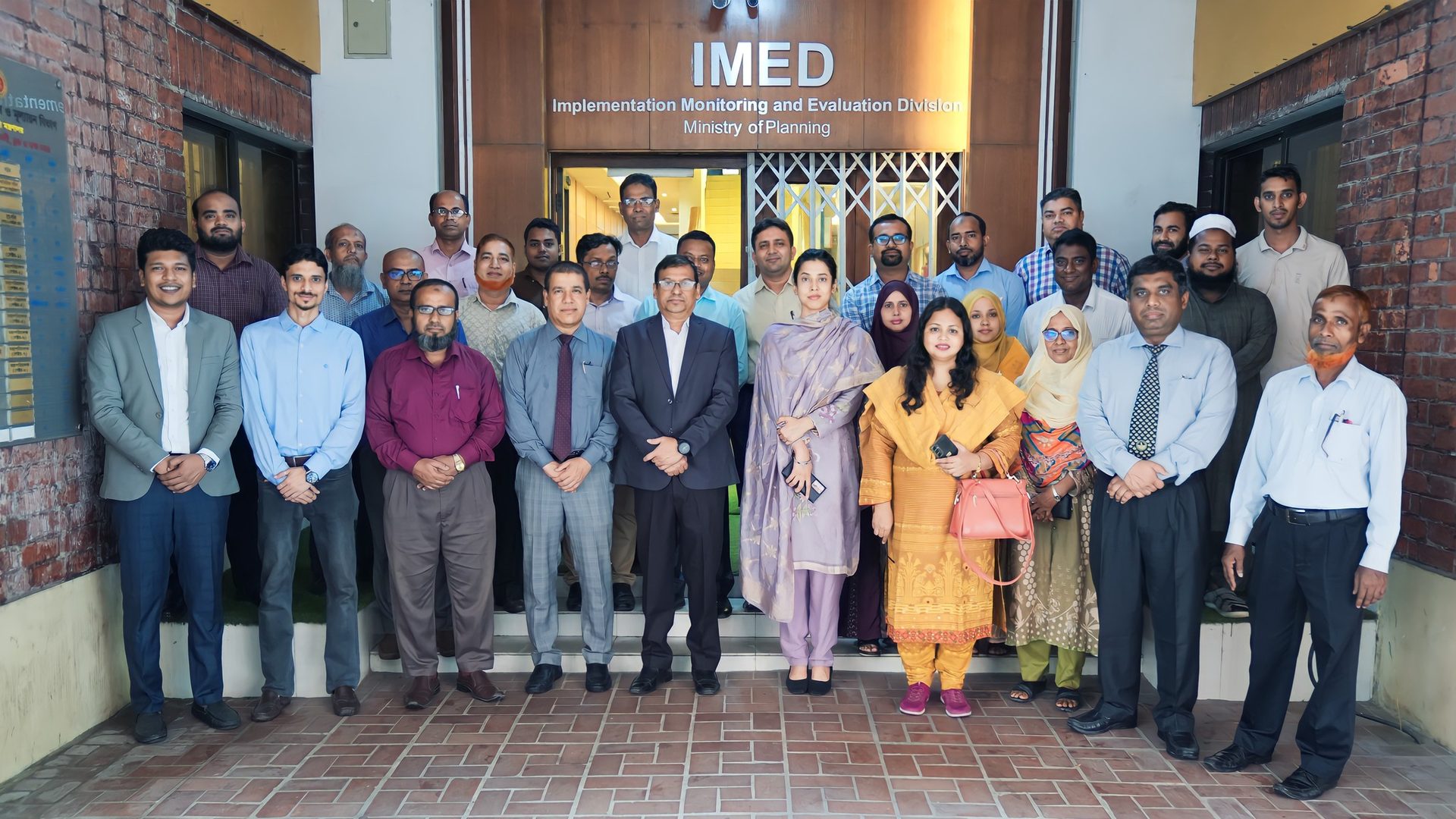 Group photo of training participants under the IMED signage, with two women in pink and lavender outfits in the front row.