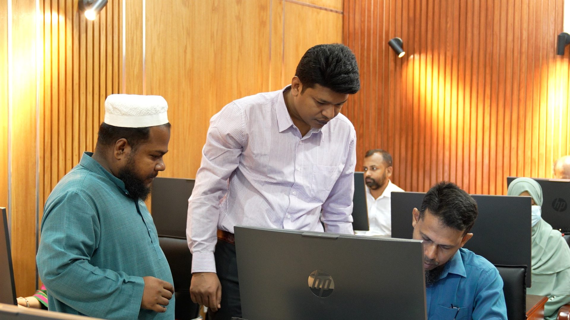 A facilitator in a maroon blazer leans over a computer to guide a seated participant wearing a white prayer cap and teal kurta.