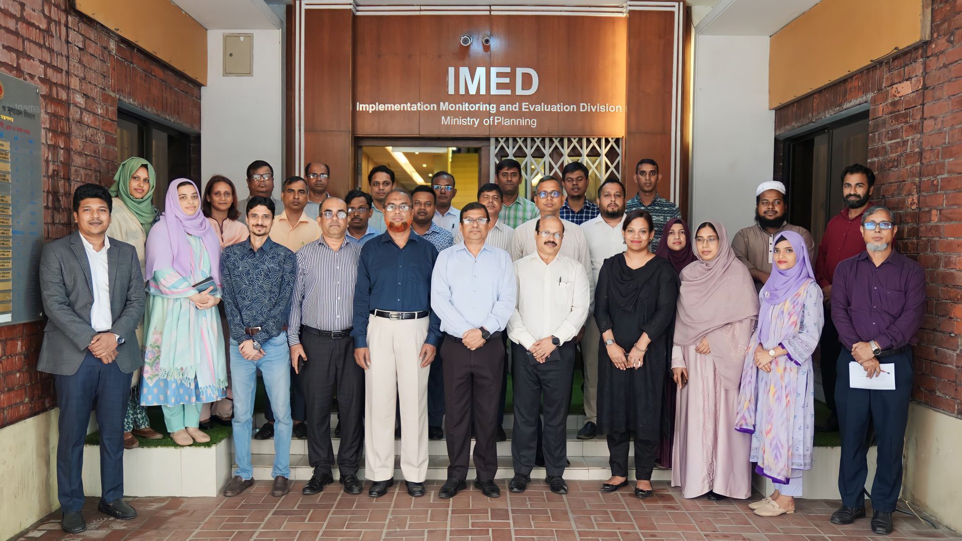 A large group photo of training participants beneath the IMED signage, with several women wearing pastel-colored shawls in the front row.