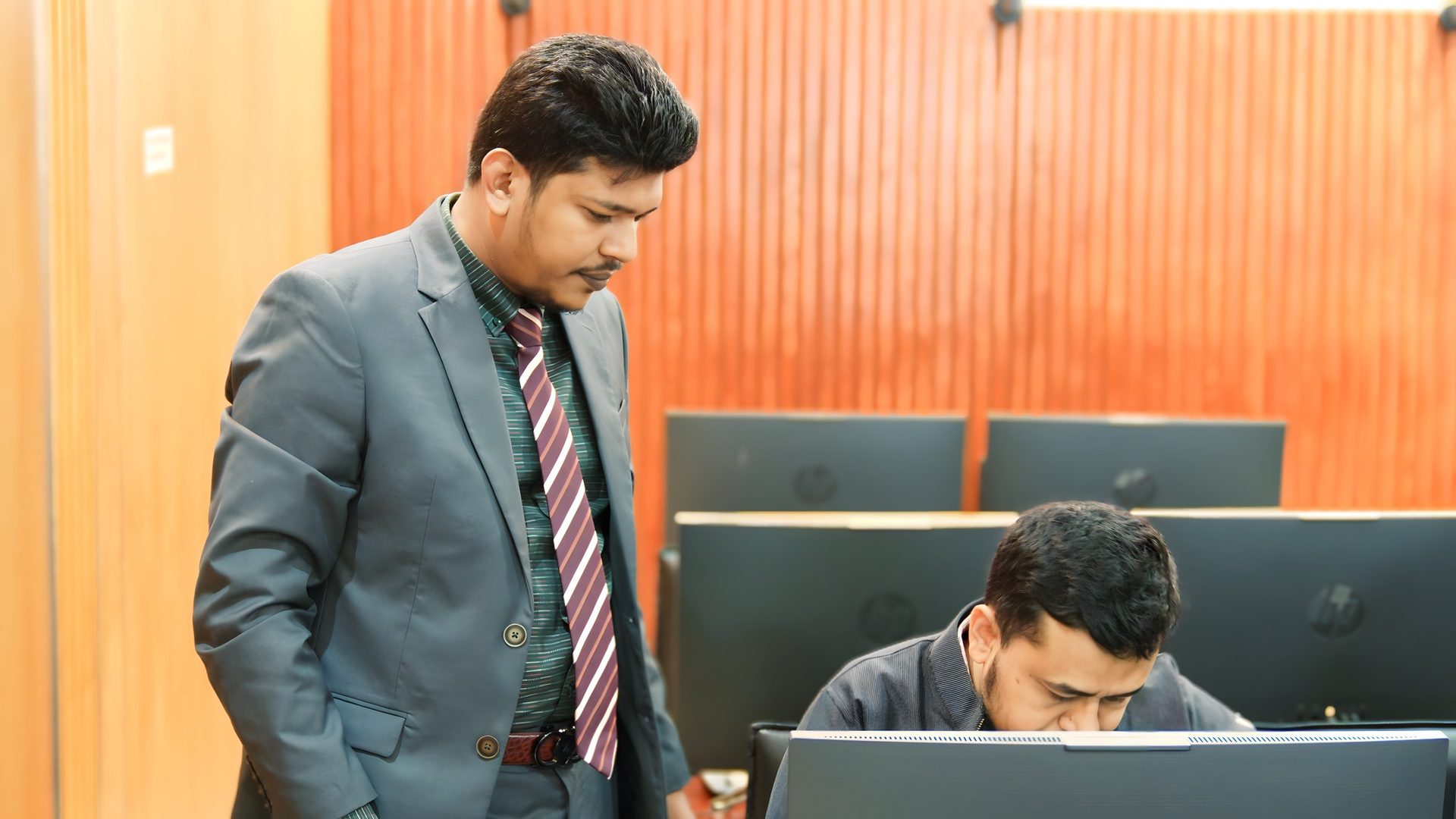 Raihan Khan stands beside a participant working at a computer station during the training session.
