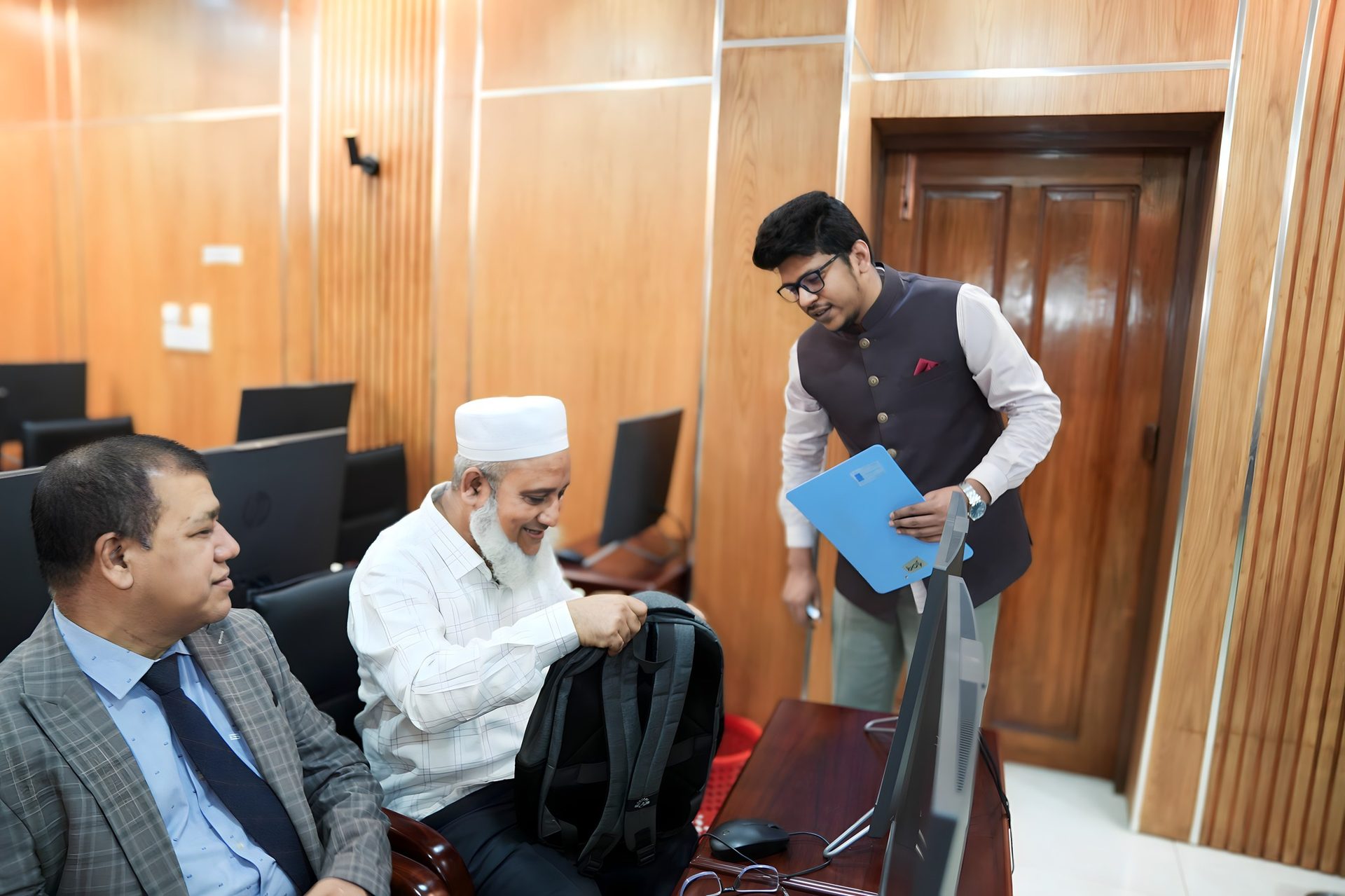 Raihan Khan, holding a folder, welcomes arriving participants including an elderly guest carrying a backpack.
