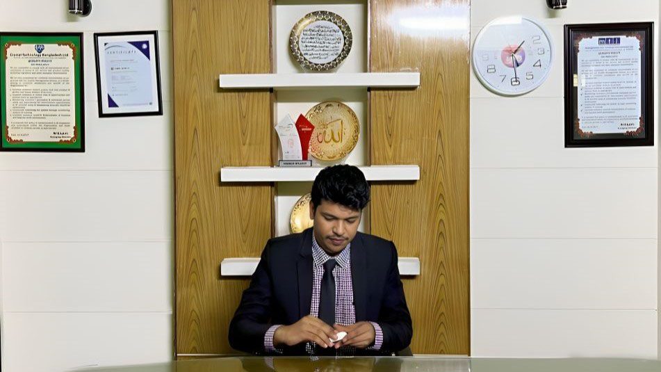 Raihan Khan seated at a reception desk in the MTI Ltd. office, surrounded by awards and certificates.
