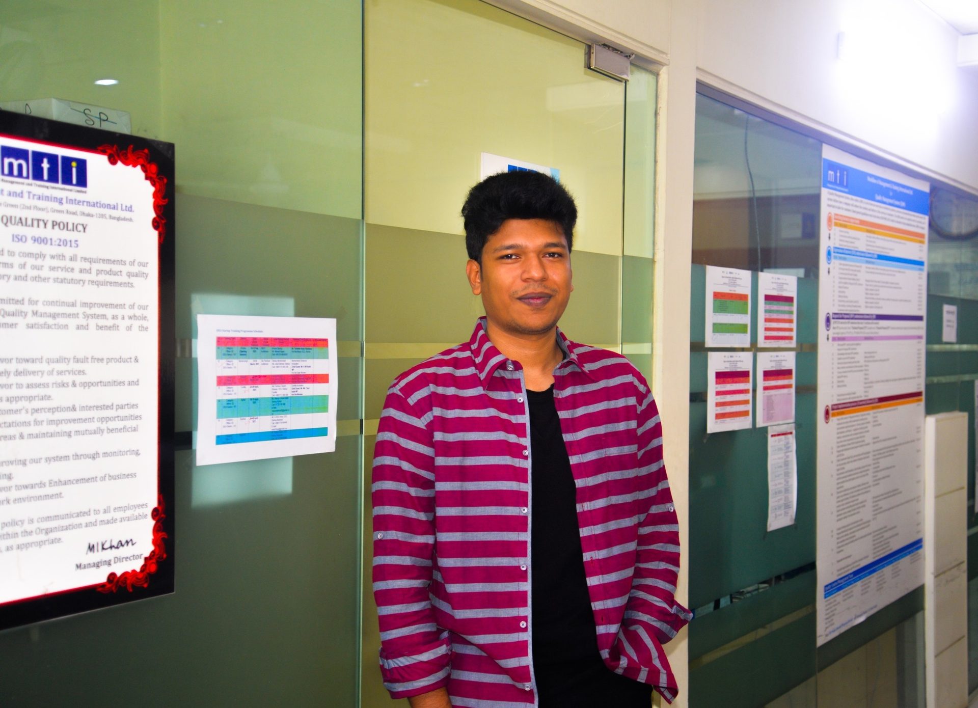 Raihan Khan standing in an MTI Ltd. office corridor beside the company's ISO 9001:2015 quality policy board.