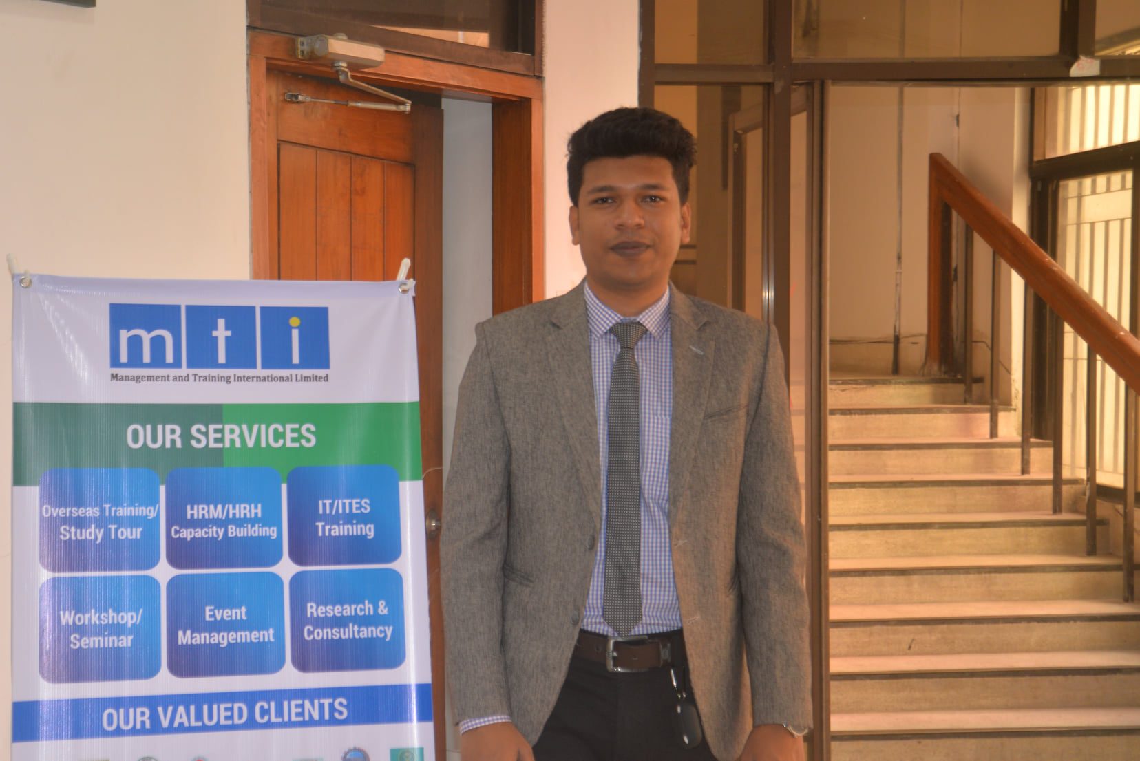Raihan Khan standing beside an MTI Ltd. services banner in the office stairwell.