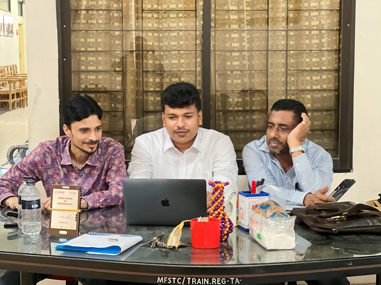 Raihan Khan seated with colleagues at a USAID-supported training program registration table.