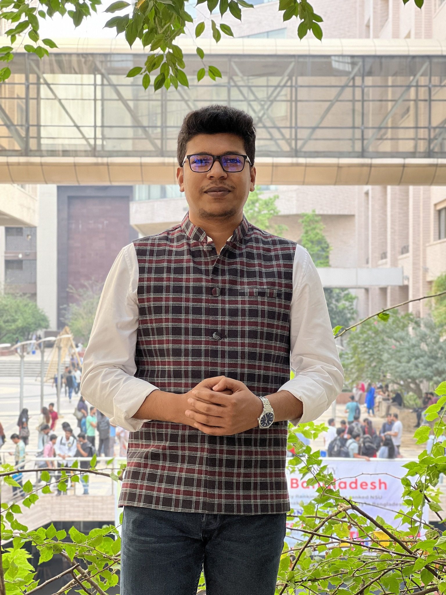 Raihan Khan in a plaid waistcoat on a campus footbridge overlooking a student gathering.
