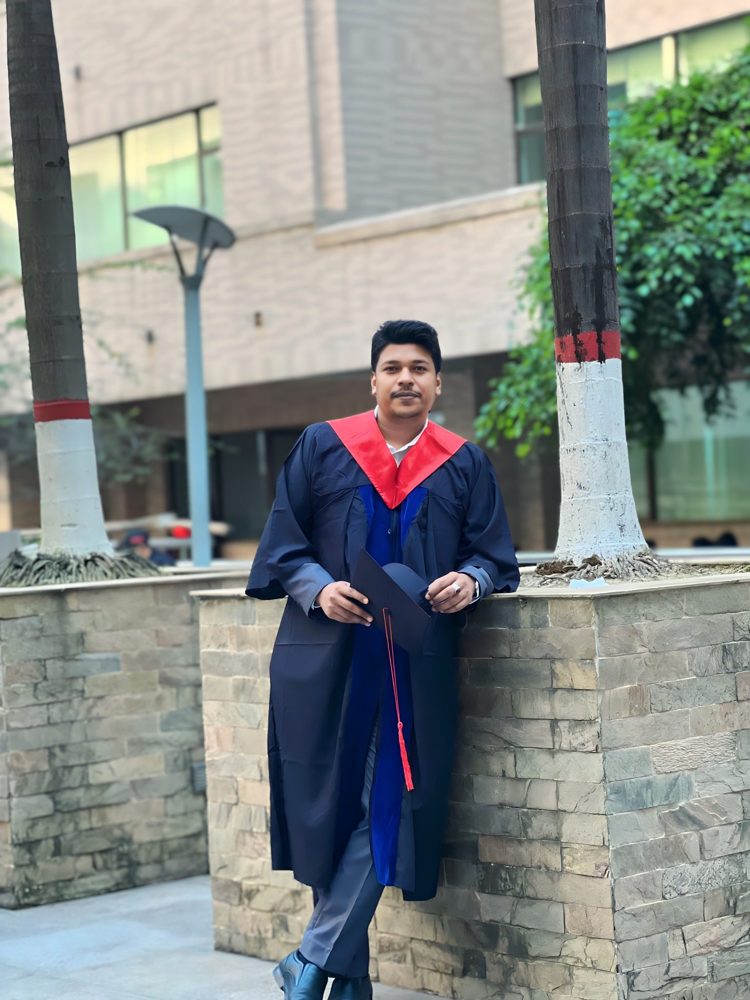 Raihan Khan in his graduation gown and cap leaning against a stone wall near the campus palm trees.