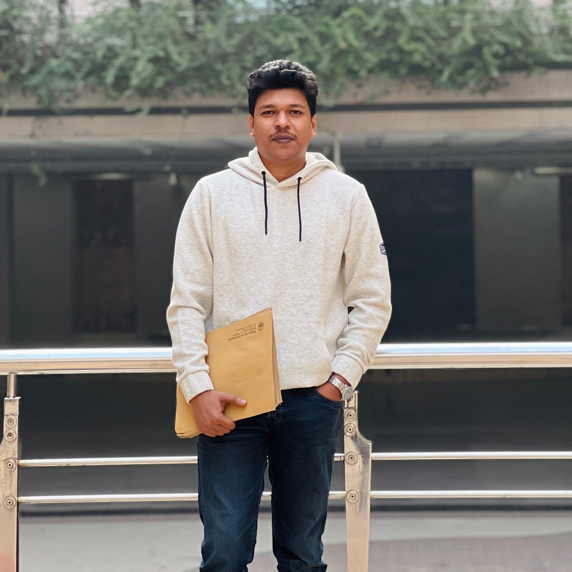 Raihan Khan in a hoodie holding university admission documents on a campus walkway.
