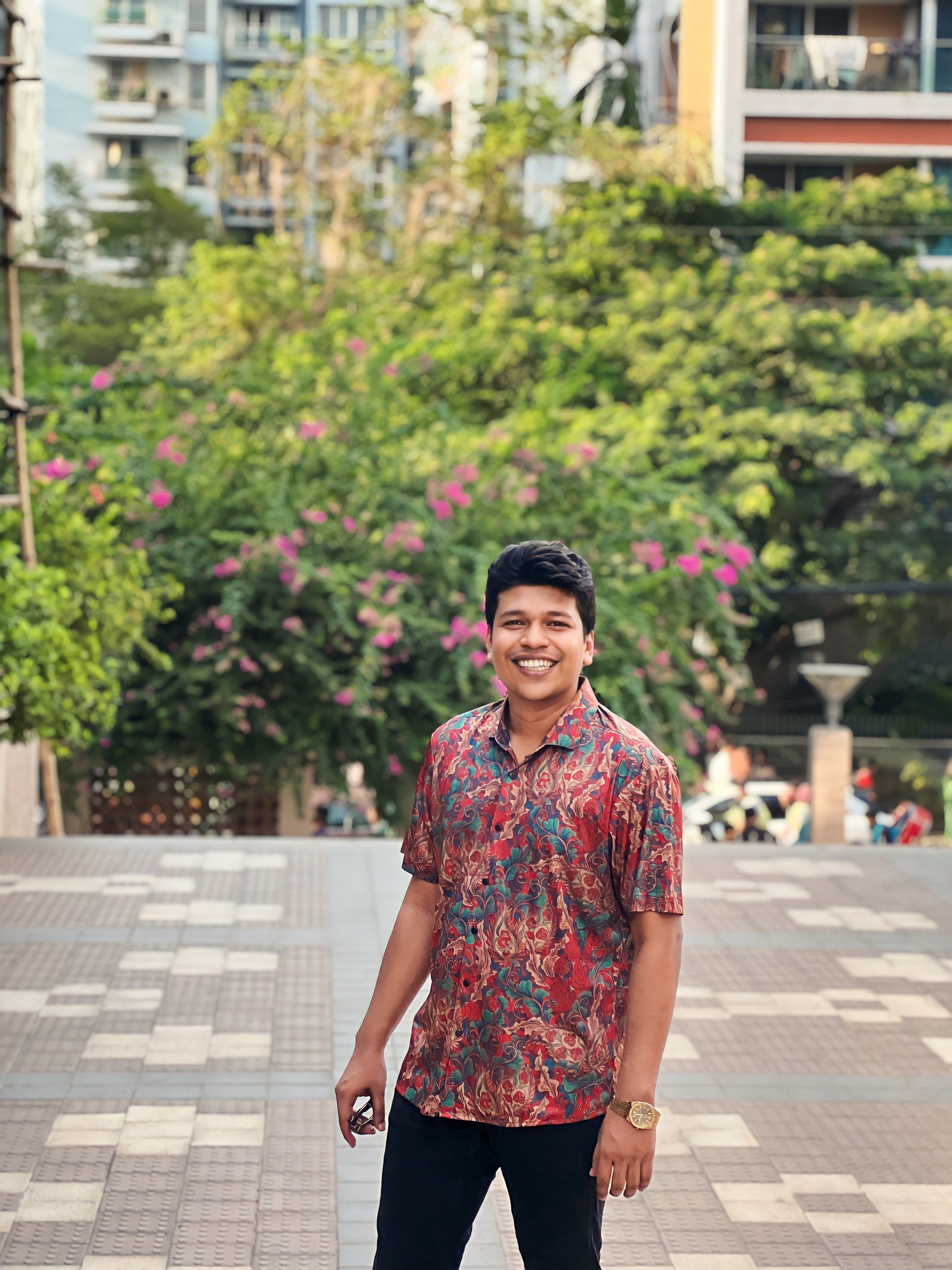 A smiling Raihan Khan in a colorful floral shirt standing in front of a flowering garden.