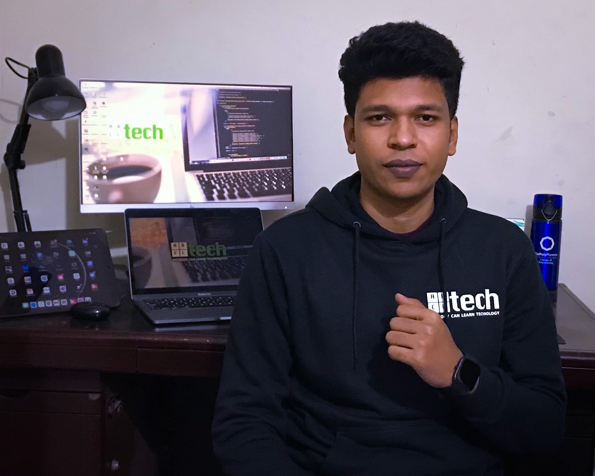 Raihan Khan in a black ABCtech hoodie seated at his desk with code visible on the laptop behind him.