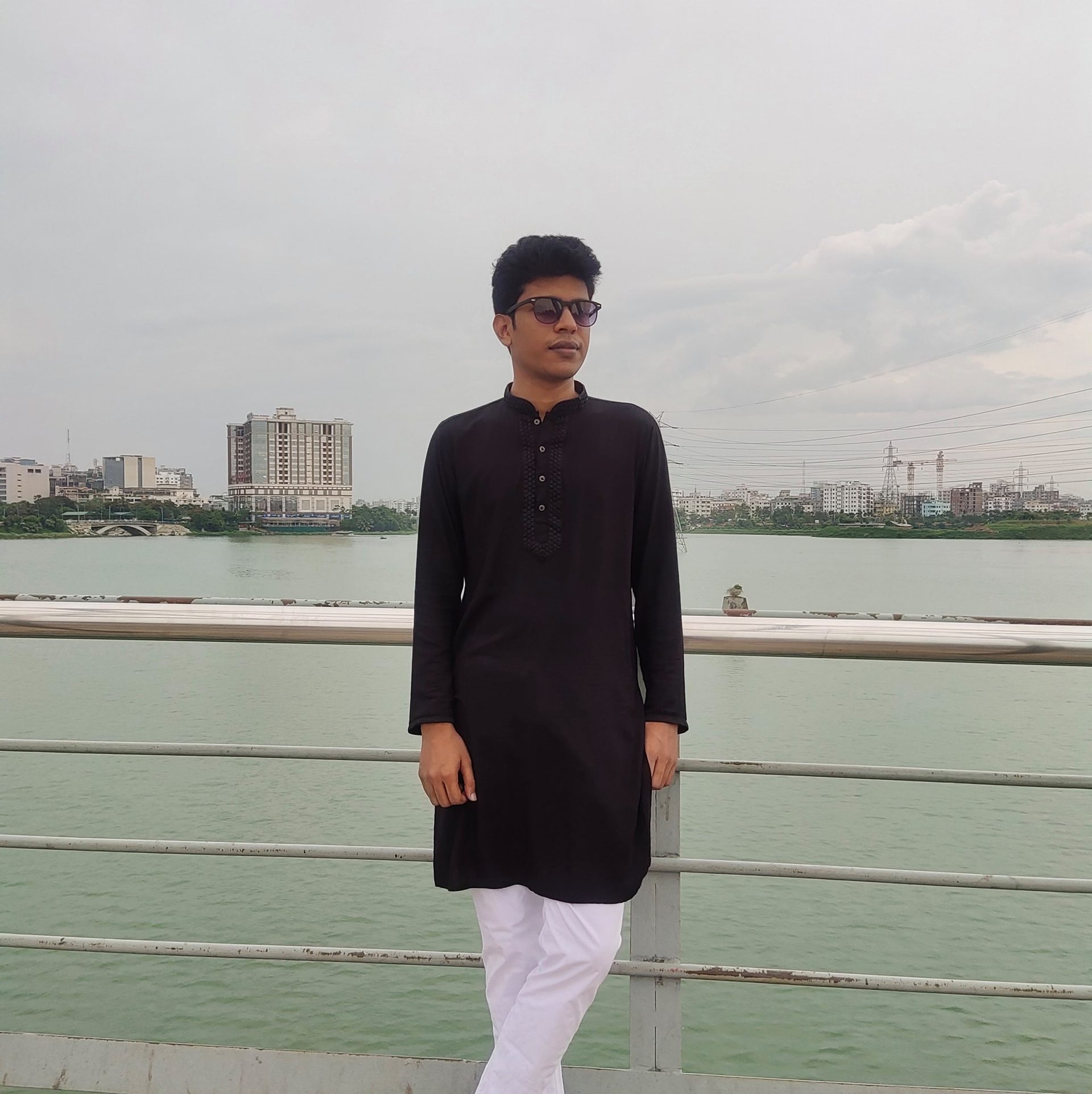 Raihan Khan in a black kurta and white pants leaning on a bridge railing above a river with the skyline beyond.