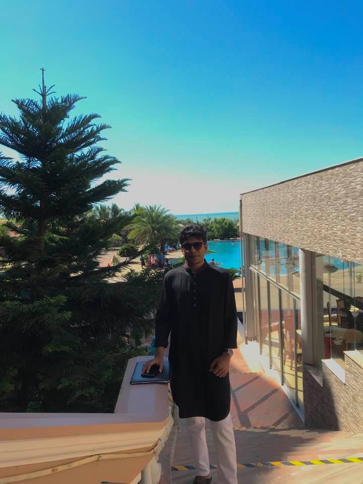 Raihan Khan in a black kurta and sunglasses standing on a resort balcony overlooking a pool and the sea.