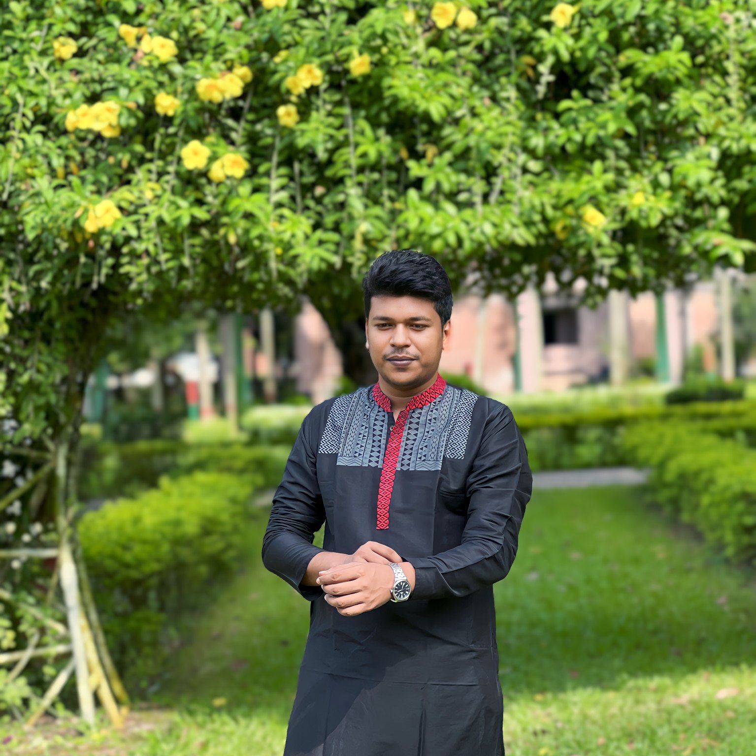 Raihan Khan in a black kurta with red trim standing beneath a tree of yellow flowers in a garden.