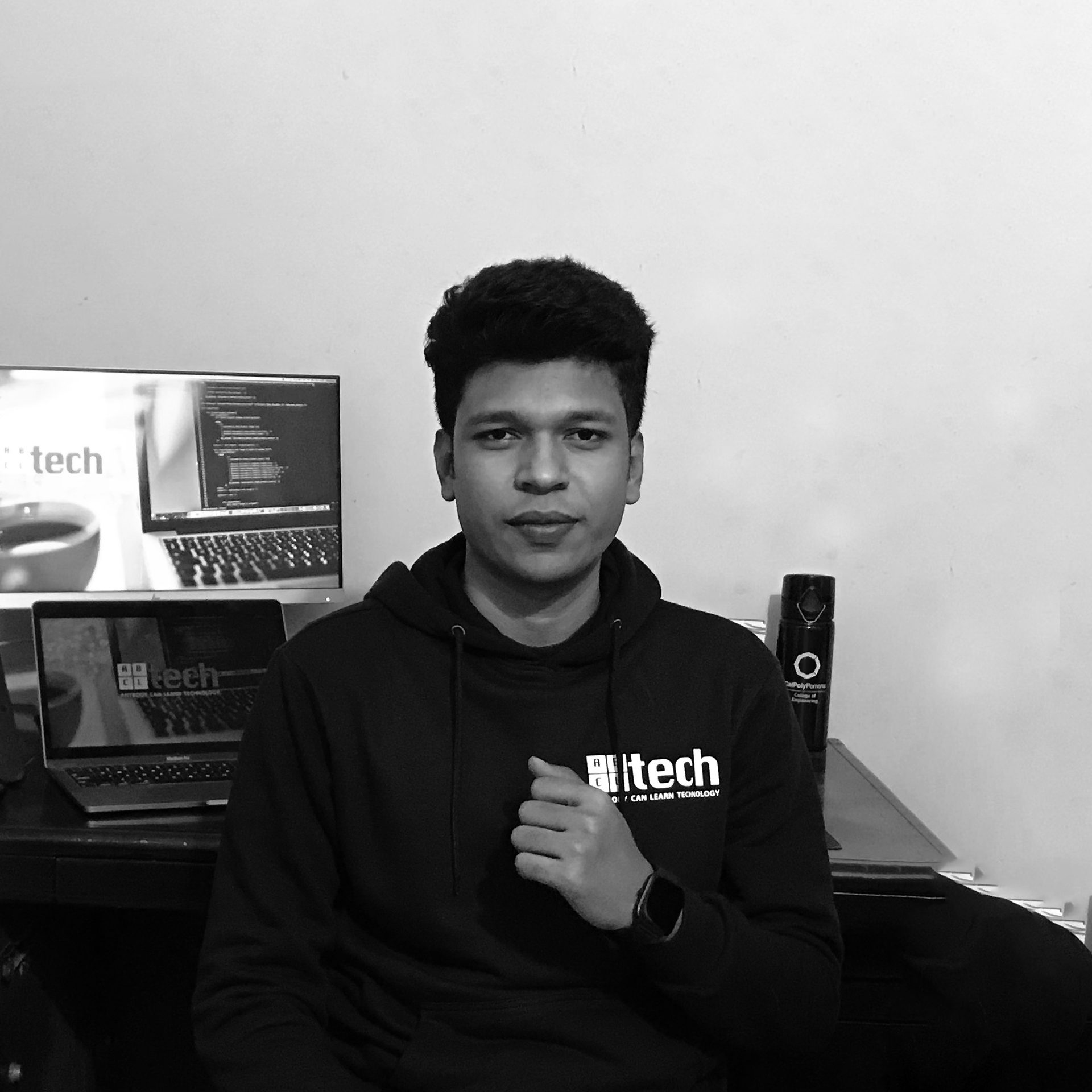 A black and white portrait of Raihan Khan in an ABCtech hoodie seated at his desk with monitors behind him.