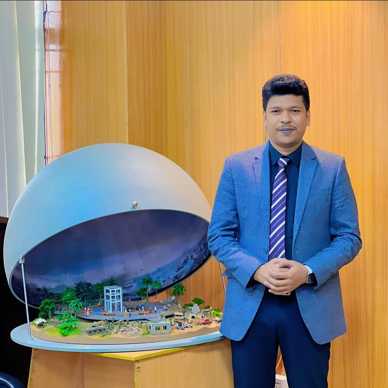 Raihan Khan in a blue blazer and striped tie standing beside a miniature diorama display in an exhibition hall.