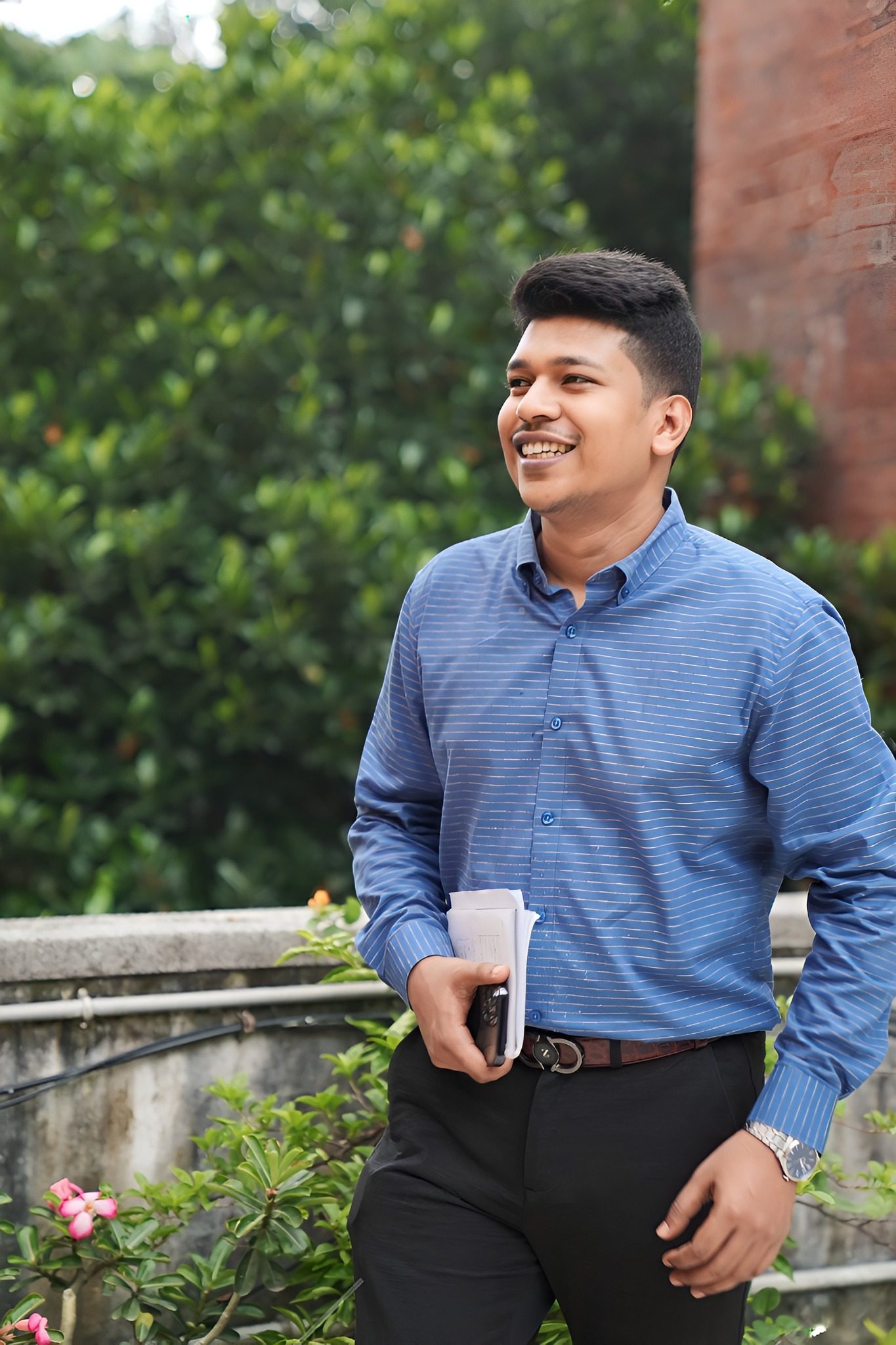 Raihan Khan in a blue striped shirt smiling while walking and holding papers and his phone outdoors.