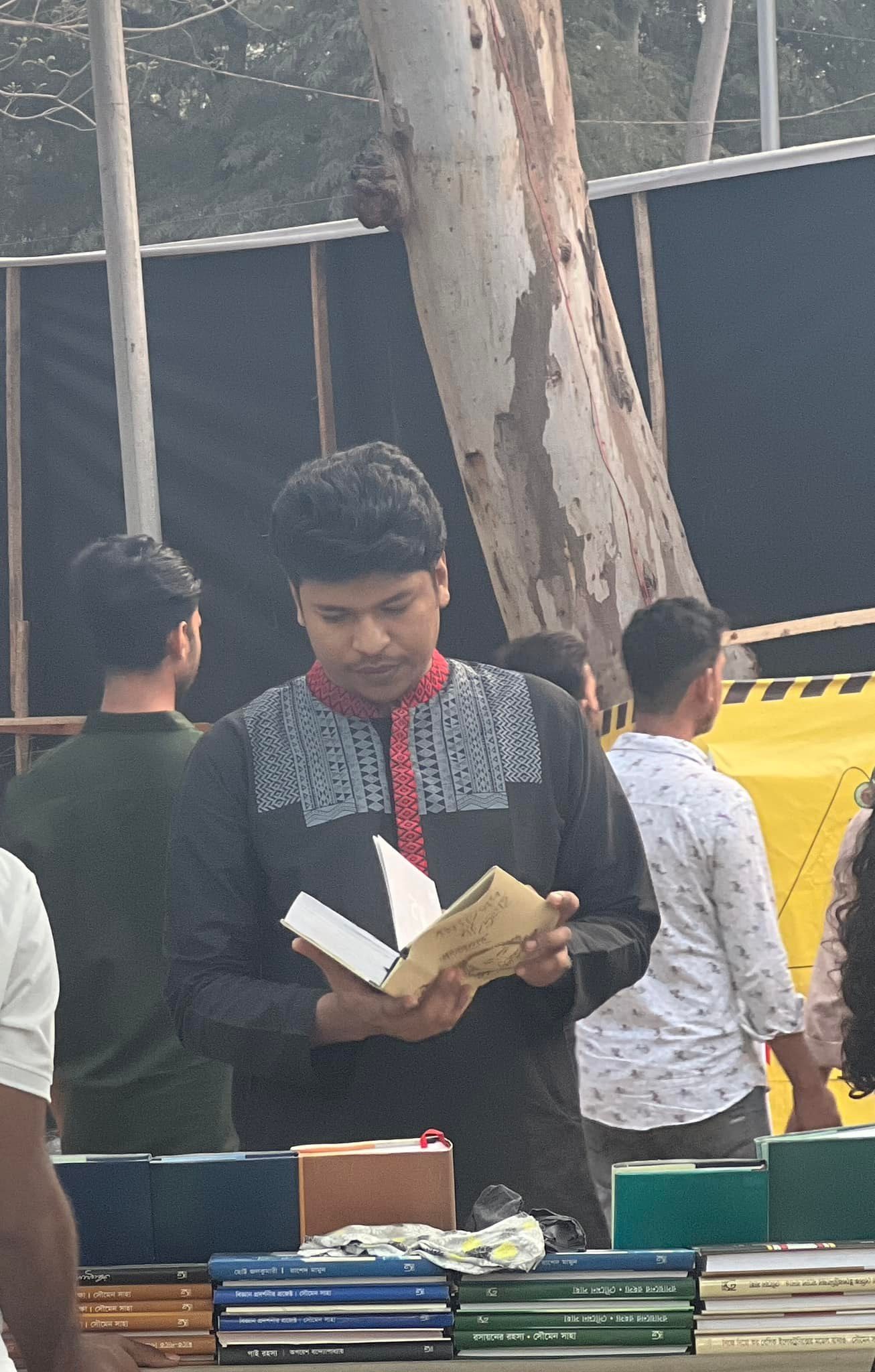 Raihan Khan in a black kurta reading a book at an outdoor book fair stall surrounded by visitors.