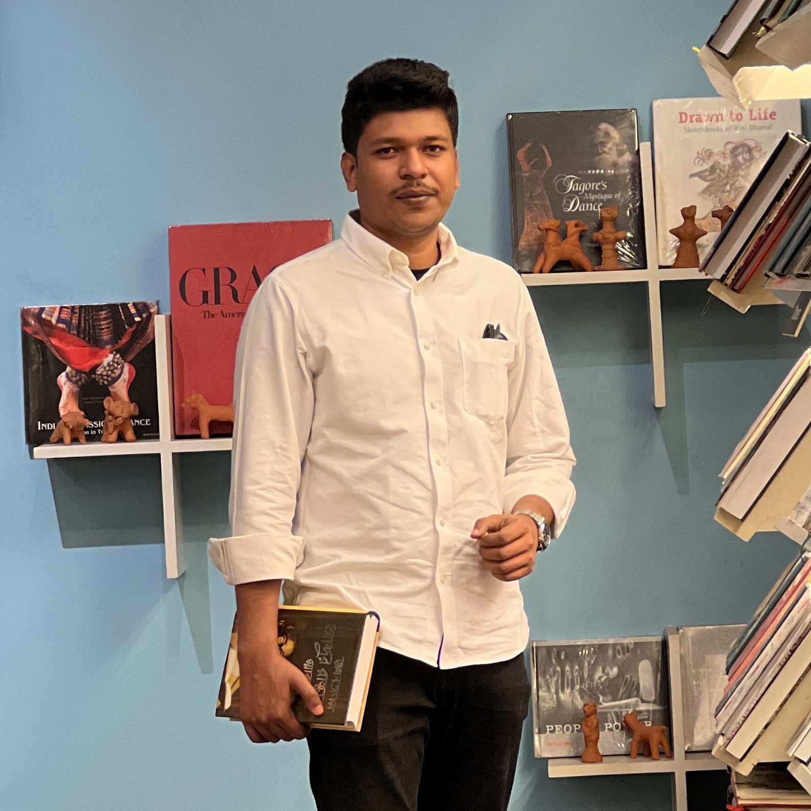 Raihan Khan in a white shirt holding a stack of books inside a bookstore with art books on the shelves behind him.