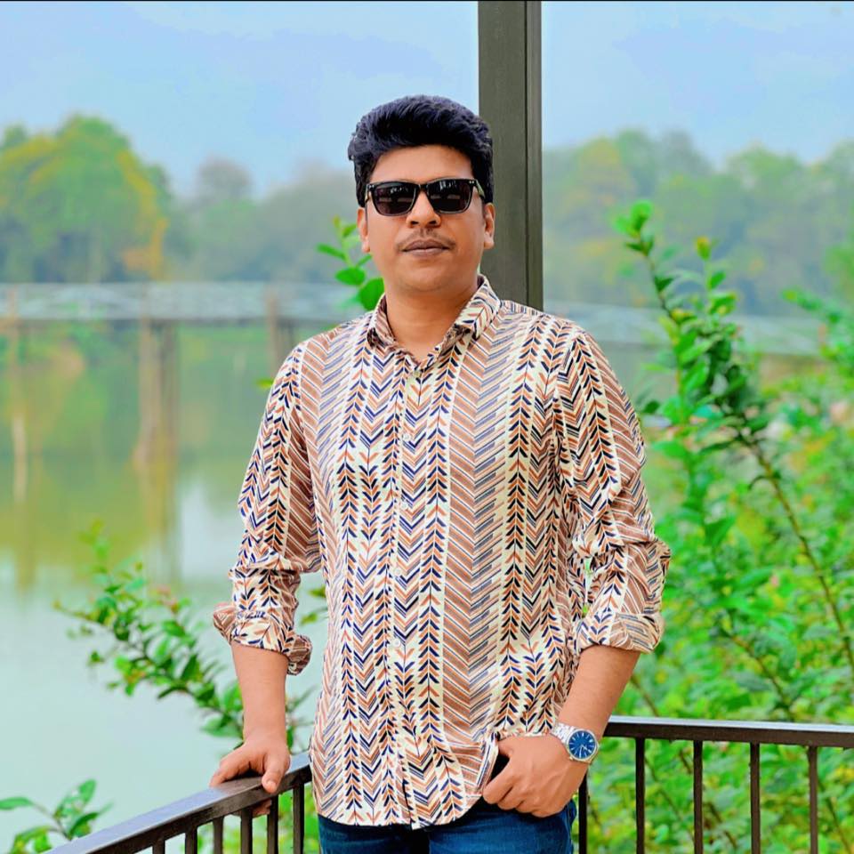 Raihan Khan in a chevron-print shirt and sunglasses resting his hand on a railing at a riverside gazebo.