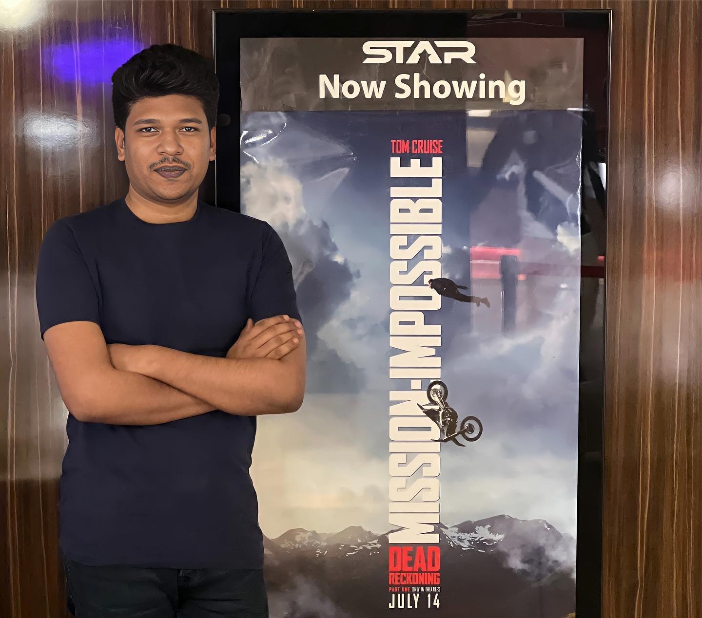 Raihan Khan with arms crossed standing beside a Mission Impossible movie poster at a cinema.