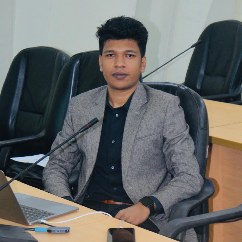 Raihan Khan in a grey blazer seated at a conference table with a microphone and laptop in a meeting room.