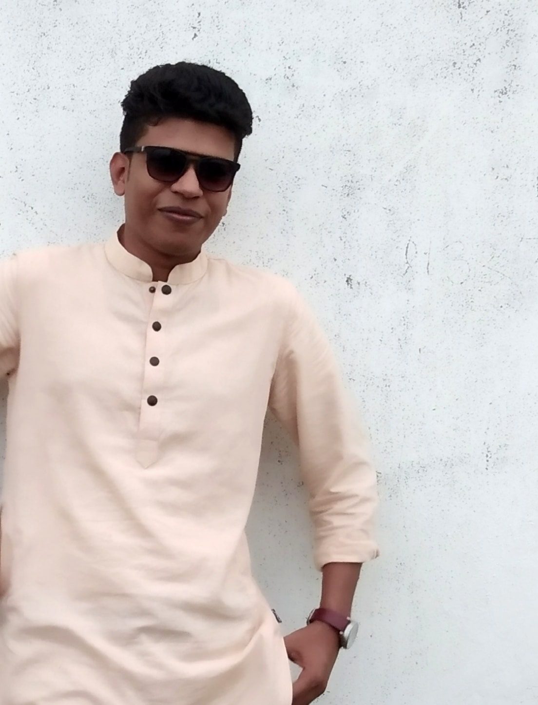 Raihan Khan in a cream kurta and sunglasses leaning against a white textured wall with a slight smile.