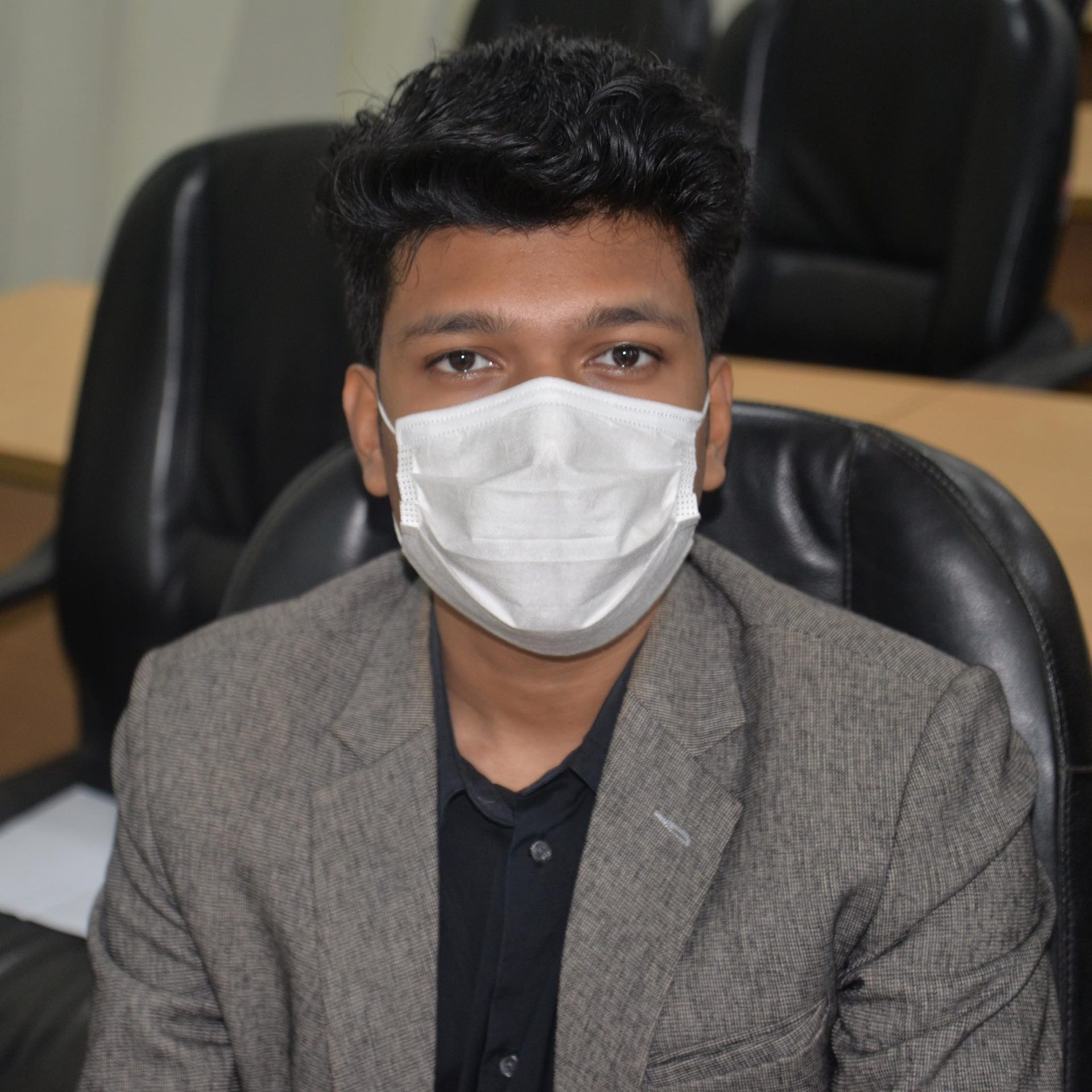 Raihan Khan in a grey blazer wearing a white face mask while seated in an office chair.