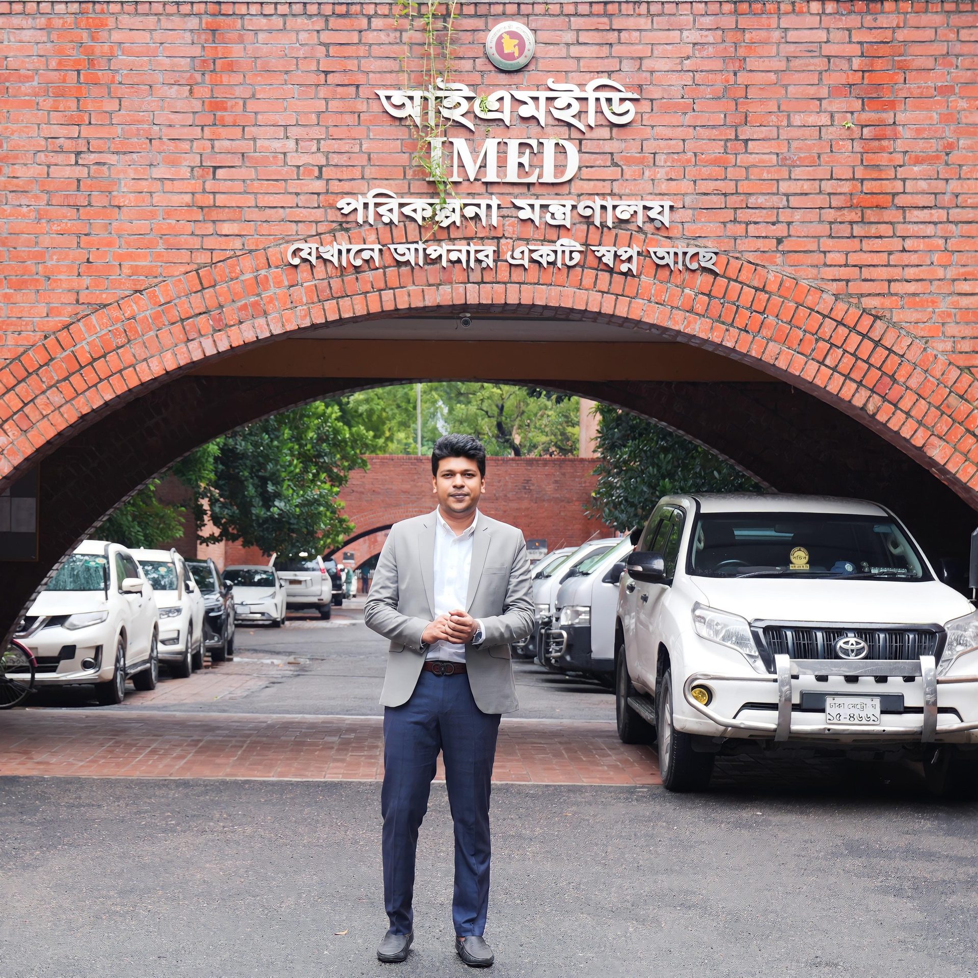 Raihan Khan in a grey blazer standing beneath a brick archway at the IMED government building entrance.