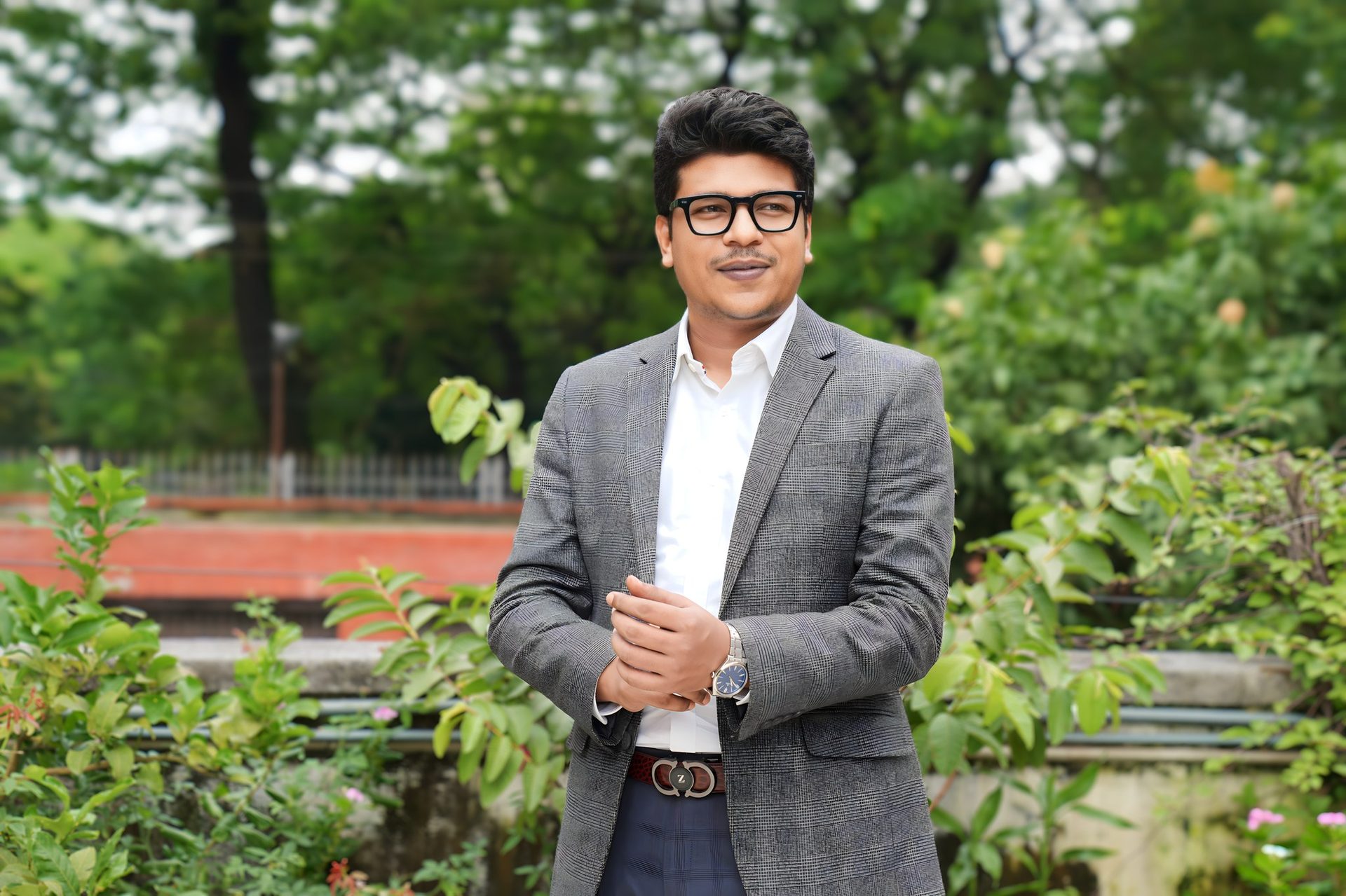 Raihan Khan in a grey plaid blazer and eyeglasses glancing to the side in a green garden.