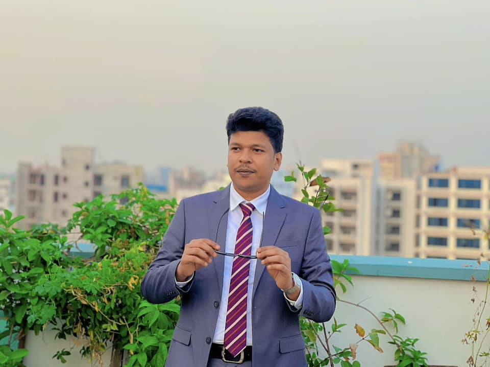 Raihan Khan in a grey suit with a purple striped tie holding his sunglasses on a rooftop overlooking the city.