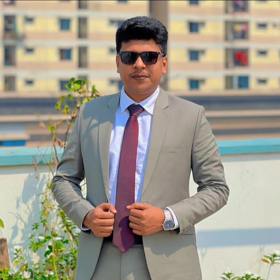Raihan Khan in a grey suit and tie adjusting his jacket on a rooftop with city buildings behind him.