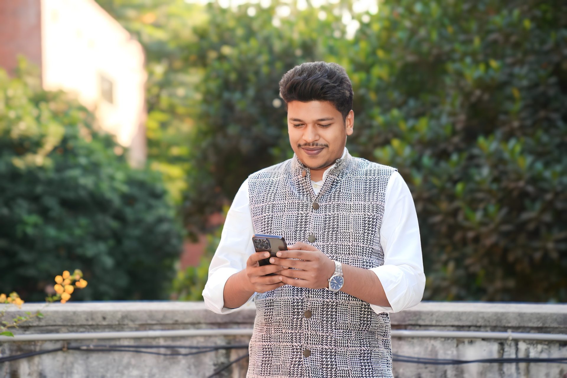 Raihan Khan in a houndstooth waistcoat over a white kurta smiling while checking his phone in a garden.