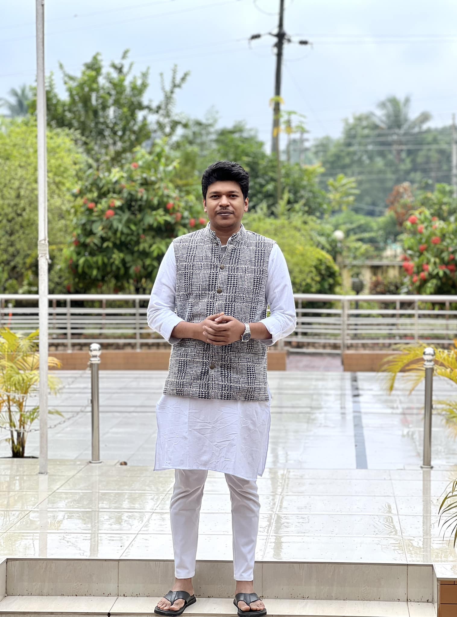 Raihan Khan in a houndstooth waistcoat over a white kurta standing on a rain-soaked plaza in sandals.