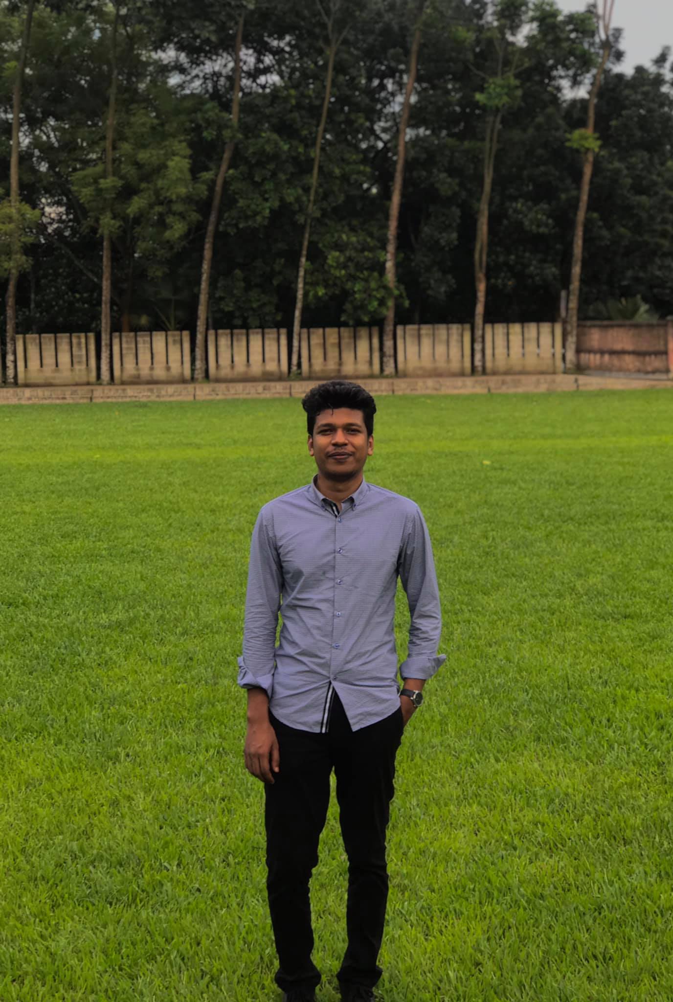 Raihan Khan in a lavender shirt standing on an open green field lined with tall trees.