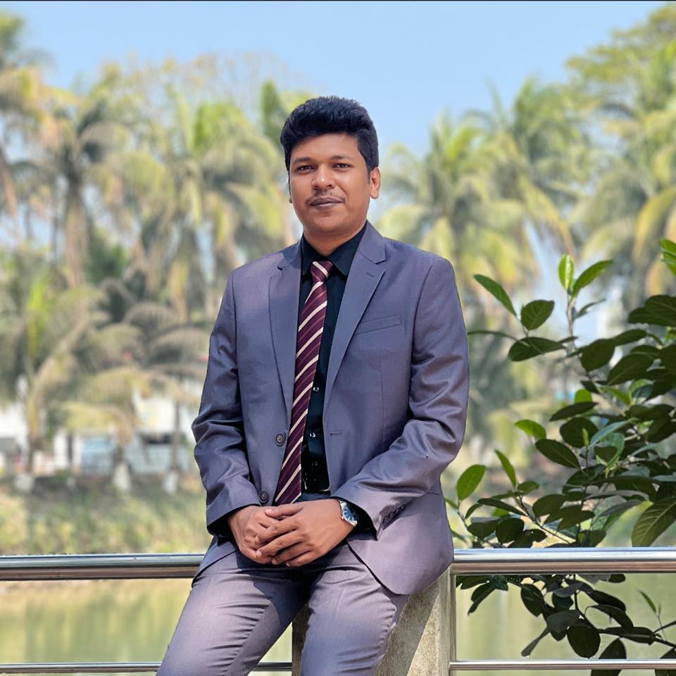 Raihan Khan in a lavender suit and striped tie sitting on a railing beside a river lined with palm trees.