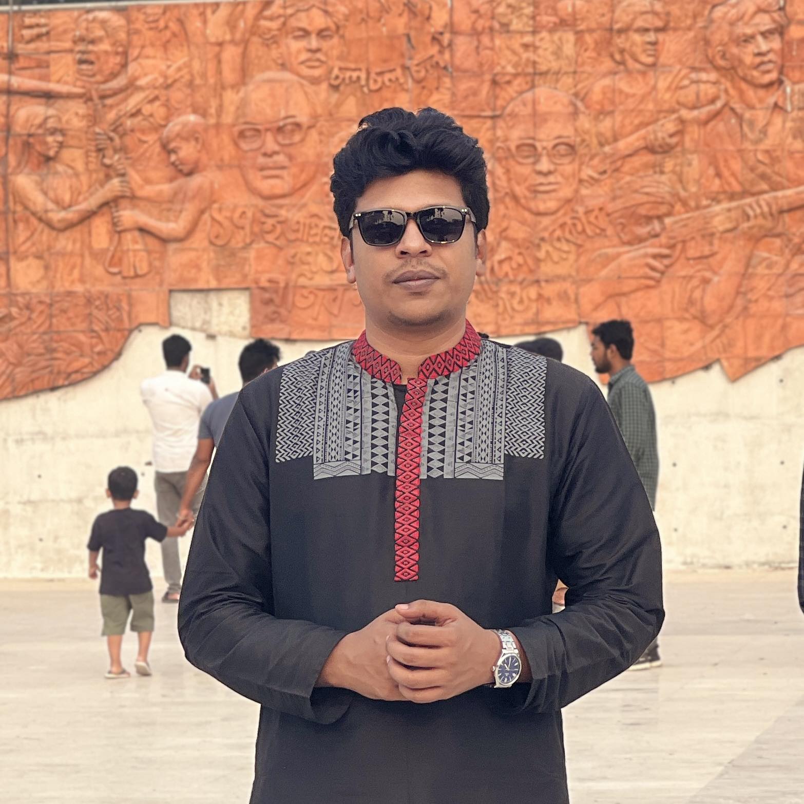 Raihan Khan in a black kurta with sunglasses standing before a relief mural depicting Bangladesh's Liberation War.