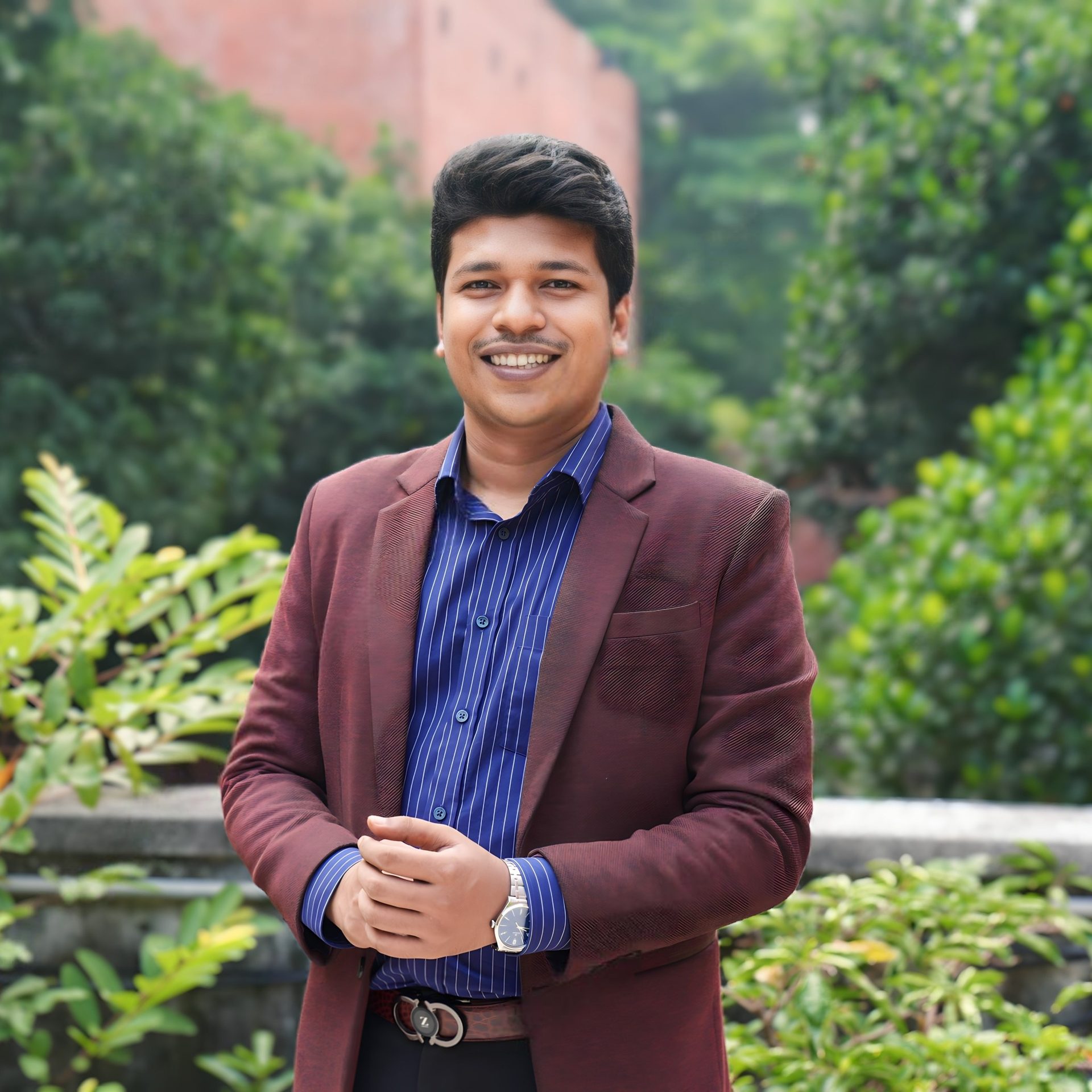 Raihan Khan in a maroon blazer and pinstriped shirt smiling broadly in front of a red brick building.