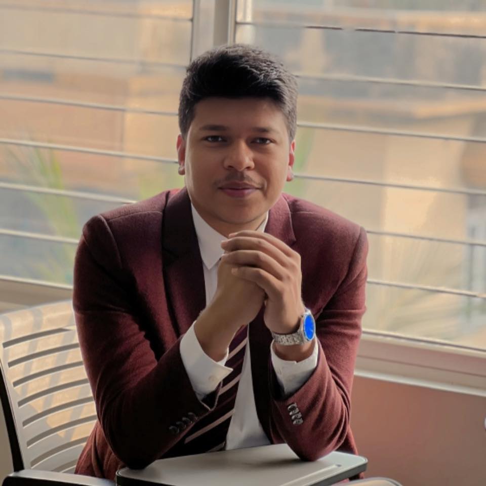 Raihan Khan in a maroon blazer and striped tie posing with clasped hands in front of window blinds.