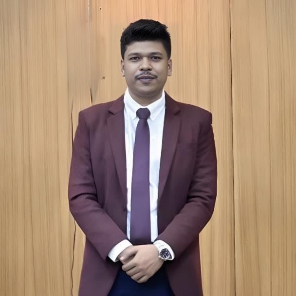 A close-up portrait of Raihan Khan in a maroon blazer and tie against a plain wooden backdrop.