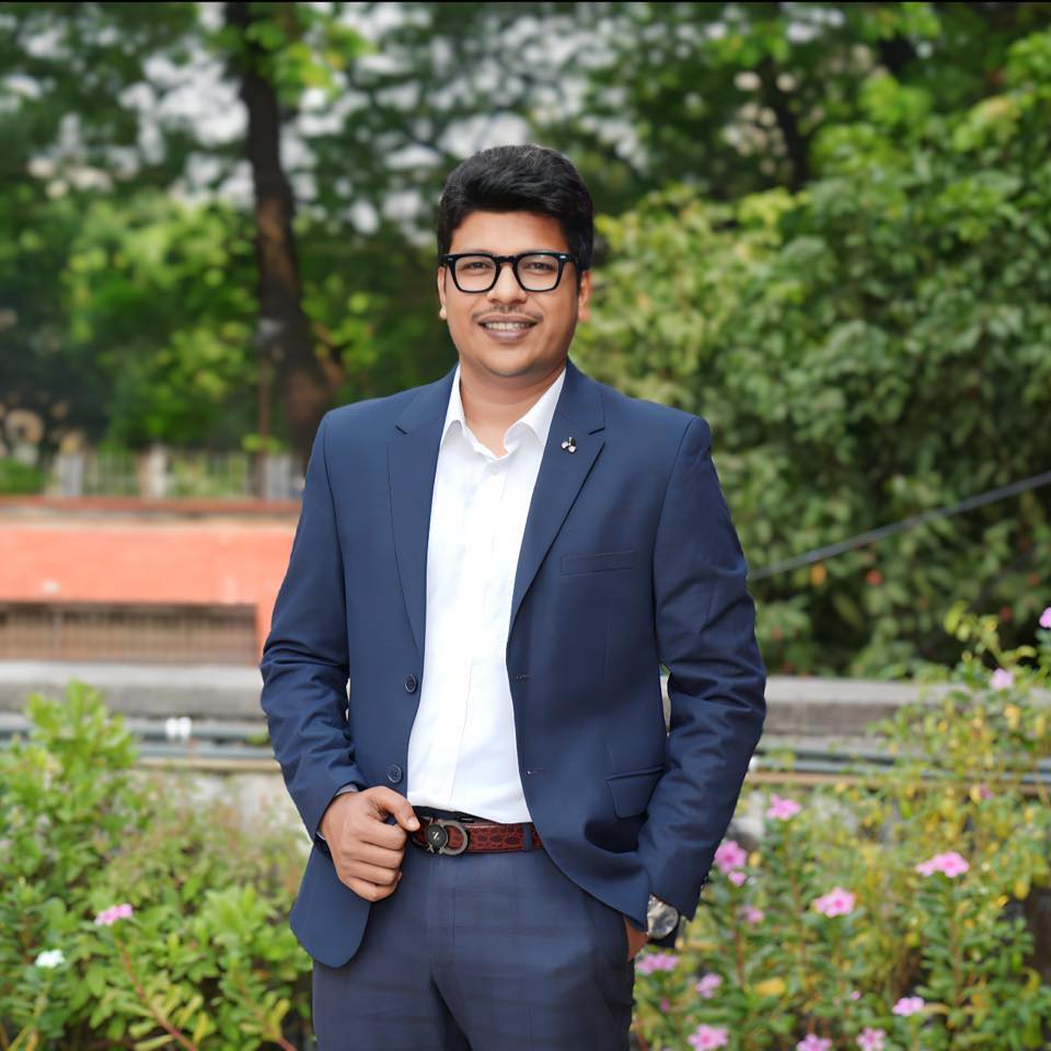 Raihan Khan in a navy blazer and eyeglasses smiling in a garden with pink flowers blooming nearby.