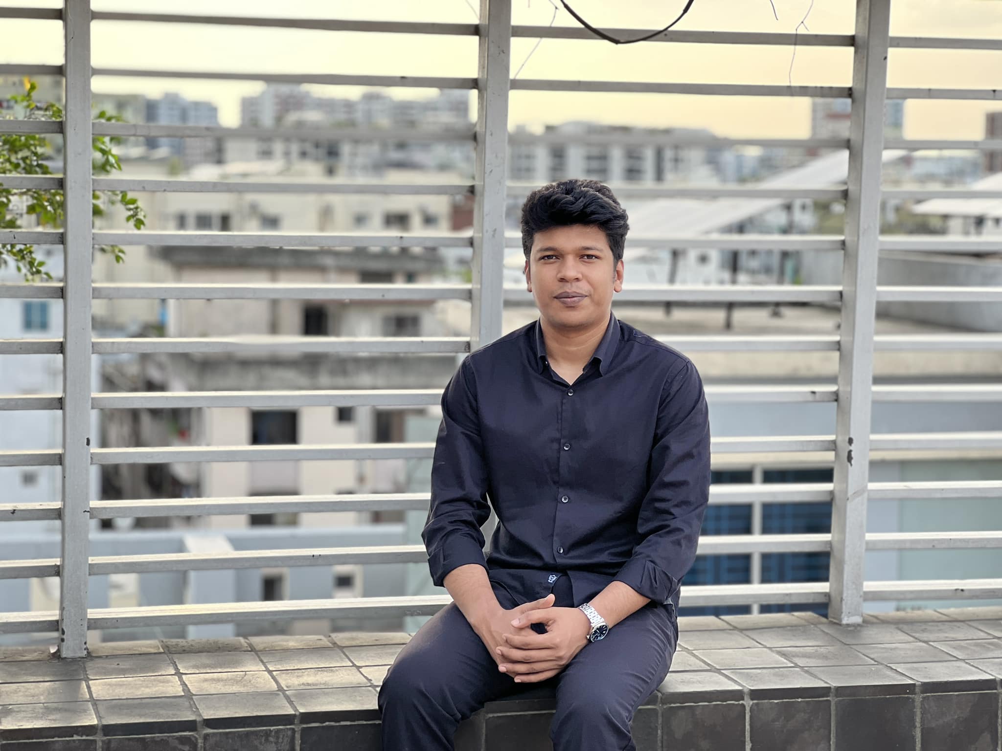 Raihan Khan in a navy shirt sitting on a rooftop ledge with city buildings in the background.